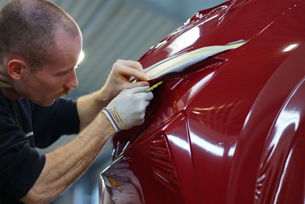 Folientechniker bei der professionellen Verarbeitung von bordeauxroter Folie am Frontspoiler eines Fahrzeugs – präzise Fahrzeugfolierung für sportliches Design und effektiven Lackschutz.